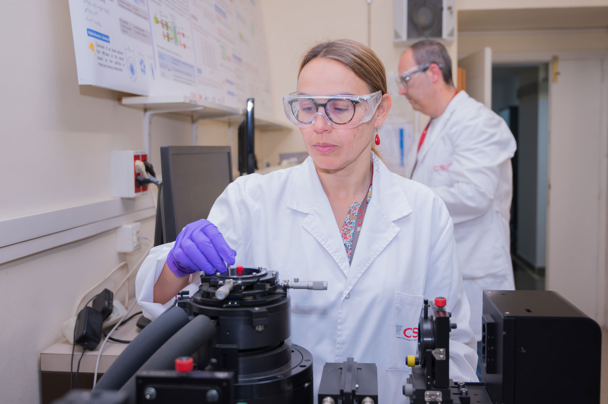 Investigadora ajusta un equipo óptico de laboratorio para estudios de química de superficies, con otro científico al fondo.